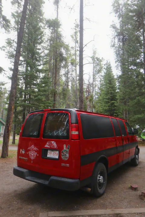 Red GMC camper van, potentially an rv rental, with adventure stickers, parked amidst tall trees in a forest setting.. Glacier Campers Vanlife Explorer 2013