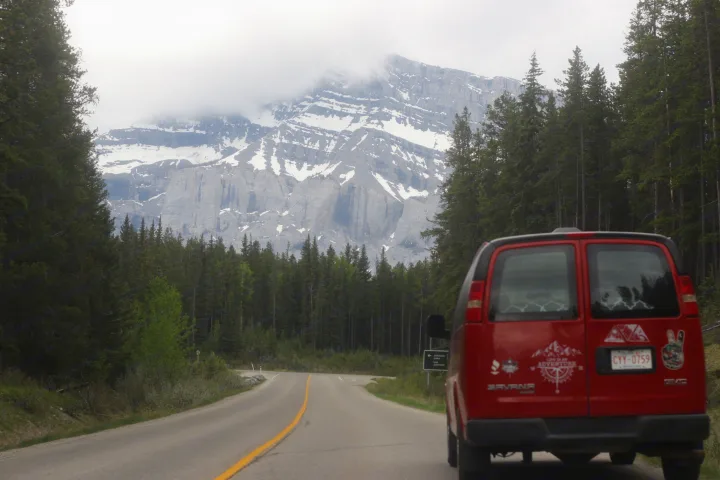 Adventure awaits! Red RV rental van drives through a forest, with massive cloud-covered, snow-capped mountains ahead.. Glacier Campers Vanlife Explorer 2013