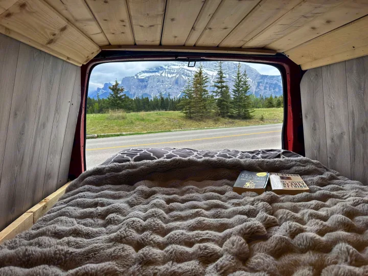 Cozy RV rental interior: A plush bed with books, offering a stunning view of snow-capped mountains & pine trees.. Glacier Campers Vanlife Explorer 2013
