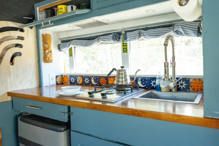 Functional RV rental kitchen featuring blue cabinets, wood countertop, stovetop, sink, and unique sun/moon tile backsplash.. Chevrolet Express s3500 2003