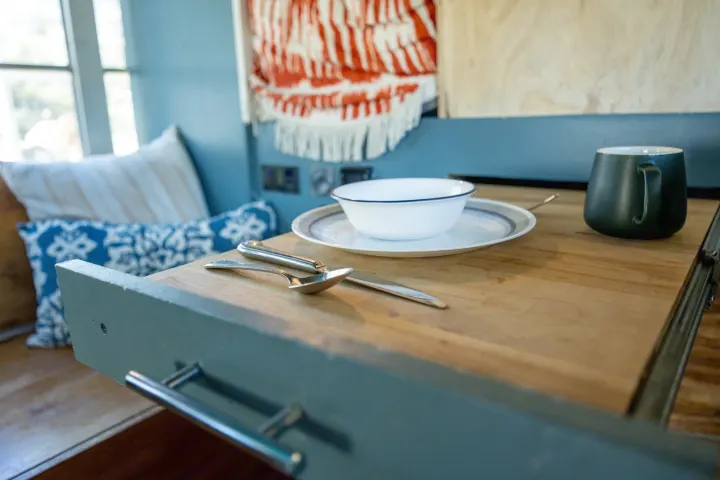 Inside a cozy rv rental: a dining table with a white bowl, plate, dark mug, and silverware, next to plush pillows.. Chevrolet Express s3500 2003