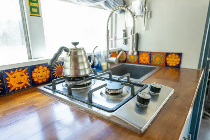 This rv rental features a bright kitchen with a stovetop, kettle, sink, and unique sun-faced tiles on a wooden counter.. Chevrolet Express s3500 2003