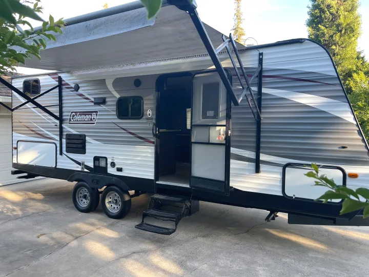This white and gray Coleman travel trailer RV rental features an extended awning, open door, and steps, parked on concrete.. Dutchmen Coleman Lantern 2018