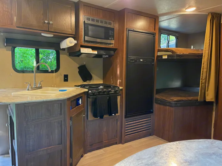 This RV rental features a full kitchen with dark wood cabinets, microwave, stove, fridge, and bunk beds with privacy curtain.. Dutchmen Coleman Lantern 2018