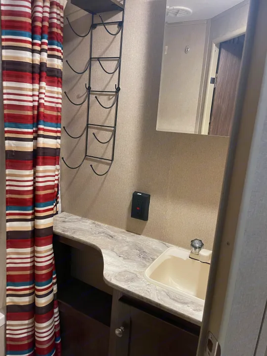 Inside an RV rental bathroom, showing a striped shower curtain, vanity with sink, mirror, and wall-mounted storage rack.. Dutchmen Coleman Lantern 2018