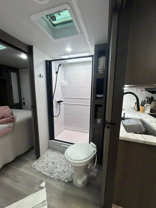 Clean RV rental with compact bathroom: white toilet, tiled shower, adjacent bedroom, and kitchen sink. Modern travel convenience.. Grand Design Transcend Xplor 2024