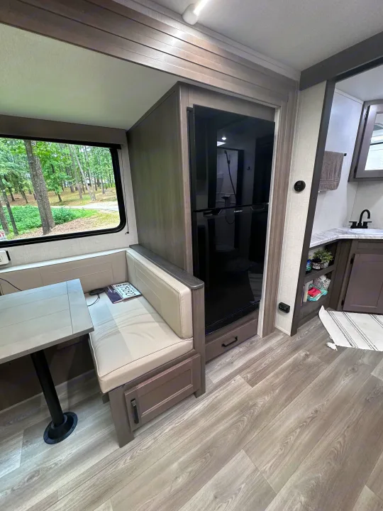 Cozy rv rental interior with a dining booth, scenic window view of trees, and a full-size fridge next to a vanity.. Grand Design Transcend Xplor 2024
