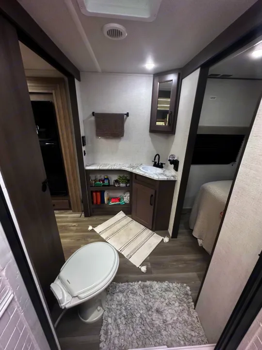 Clean RV rental bathroom features a toilet, sink, storage, and cozy rugs, with glimpses of the bedroom and living area.. Grand Design Transcend Xplor 2024