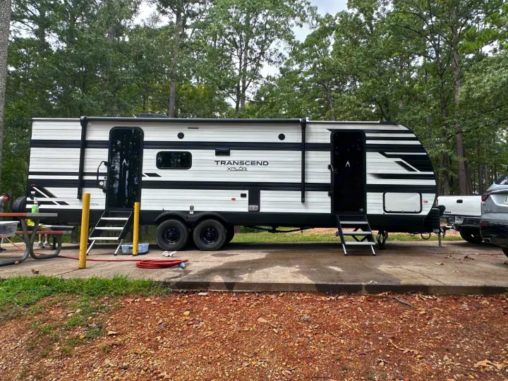This RV rental is a white and black travel trailer, parked at a wooded campsite with a picnic table.. Grand Design Transcend Xplor 2024