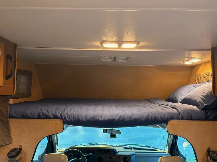 Inside an rv rental: a cozy cab-over bunk bed with blue bedding, overlooking the driver's area and windshield.. Thor Majestic 2020