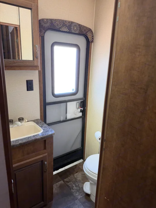 Inside an RV rental bathroom with a dark wood vanity, light sink, toilet, and an exterior door with a bright window.. Crossroads RV Rezerve 2015