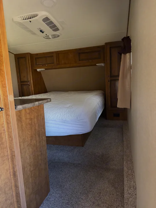 Cozy RV rental bedroom featuring a queen bed with white sheets, wooden cabinets, an overhead AC unit, and carpeted floor.. Crossroads RV Rezerve 2015