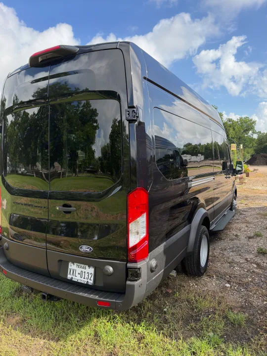 Black Ford Transit van for RV rental, detailed from the rear, reflecting sky & trees on its sleek surface.. Ford Other 2026