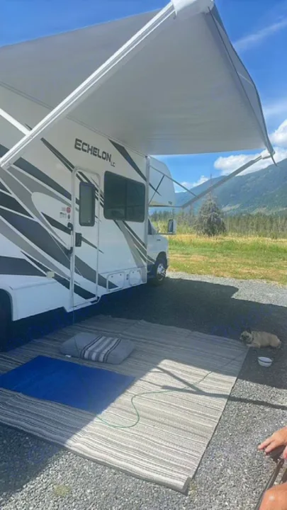 An Echelon LC RV rental under a sunny sky. Its awning shades an outdoor mat with a blue cushion and a pug, mountains in background.. Thor Echelon 2022