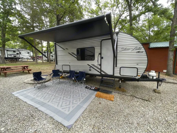 Enjoying an RV rental setup! A travel trailer with an extended awning, outdoor rug, and chairs at a wooded campsite.. East to West Silver Lake 2023