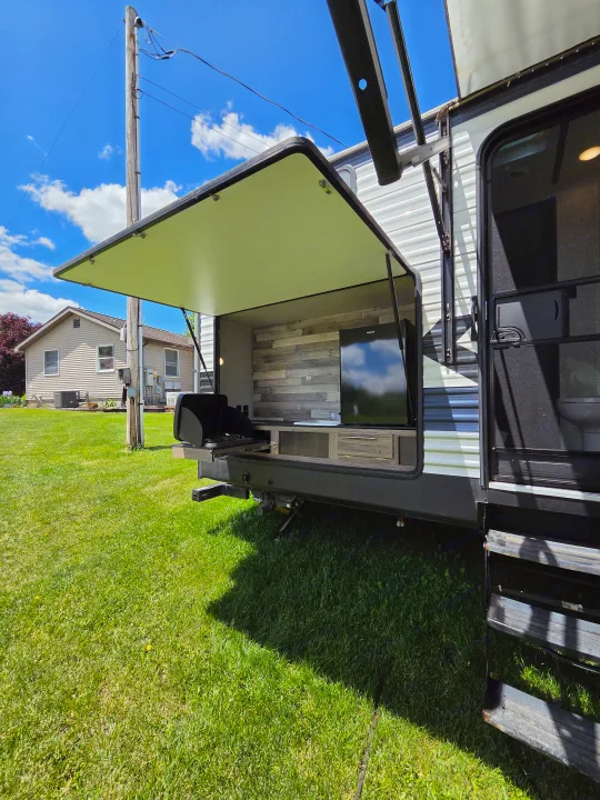 An RV rental's exterior, showing an open outdoor kitchen with a grill, TV, and counter space in a grassy area.. Crossroads RV Zinger 2021