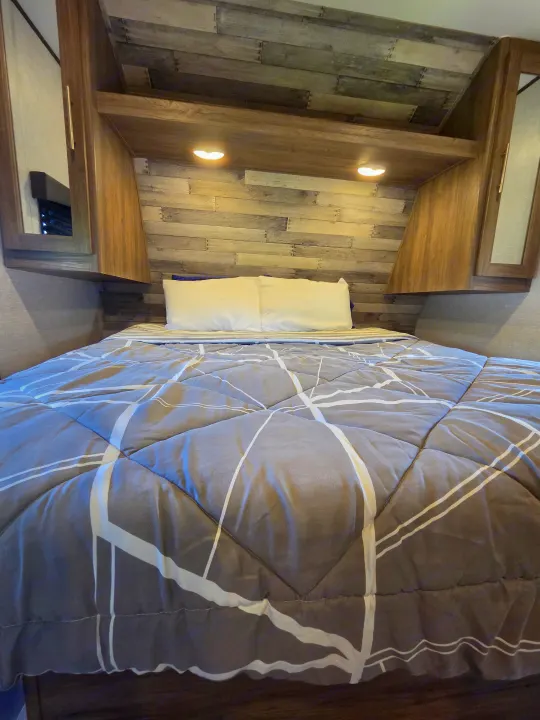 Cozy RV rental bedroom with a comfortable bed, gray geometric comforter, white pillows, wood paneling, and warm lighting.. Crossroads RV Zinger 2021