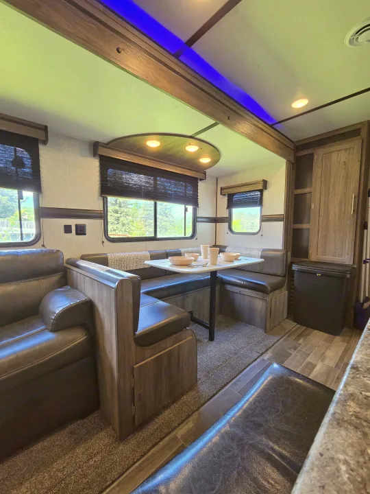 RV rental interior: A cozy dinette with dark leather seats, sofa, wood accents, and blue ceiling lights, offering outdoor views.. Crossroads RV Zinger 2021
