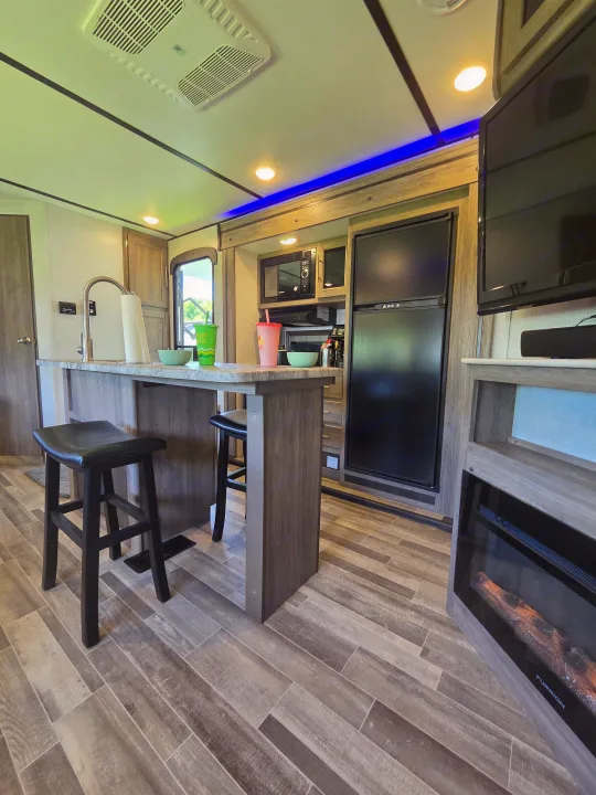 Bright RV rental interior: kitchen bar with stools, sink, fridge, microwave, and a living area featuring a TV and electric fireplace.. Crossroads RV Zinger 2021