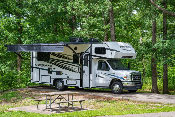 White Sunseeker RV rental parked at a wooded campsite with its awning out, next to a picnic table.. Forest River Sunseeker LE 2024