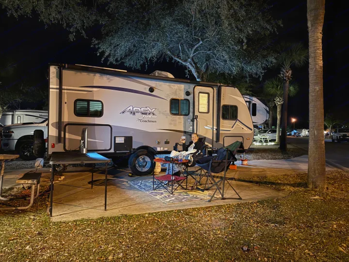 Nighttime relaxation at an RV rental campsite. A person enjoys outdoor living by their illuminated travel trailer.. Coachmen Apex Nano 2022