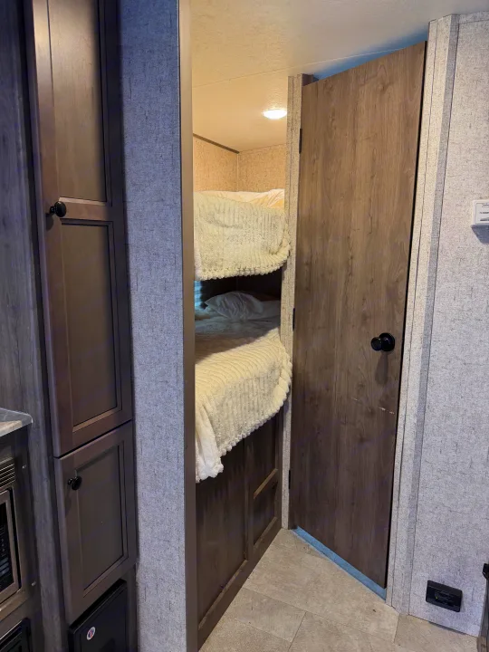 RV rental interior: A peek at cozy bunk beds with white bedding through a doorway, flanked by wood-look storage and a closed door.. Coachmen Apex Nano 2022