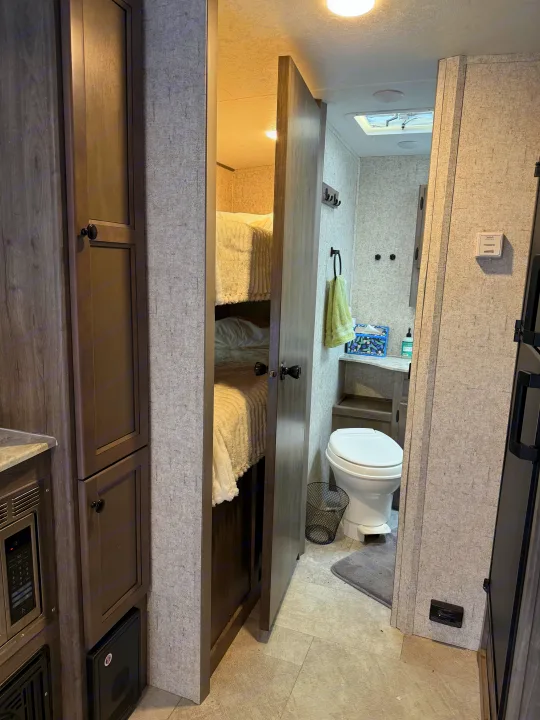 RV rental interior: narrow view of a compact bathroom with toilet, sink, and an adjacent bunk bed area, showcasing efficient space.. Coachmen Apex Nano 2022