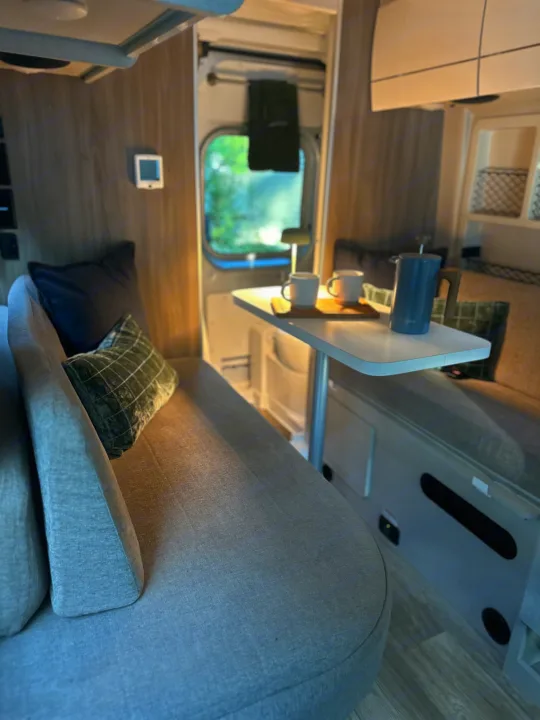 Cozy RV rental interior featuring a sofa with pillows, a dining table with coffee, and a view of nature for travel adventures.. Thor Motor Coach Rize 18G pop top 2024