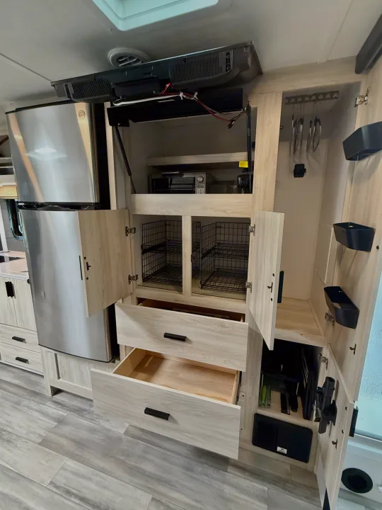 Compact RV rental kitchen/pantry. Features stainless steel fridge, light wood cabinets, open drawers, an overhead TV, and wire basket storage.. Forest River Sunseeker MBS 2022