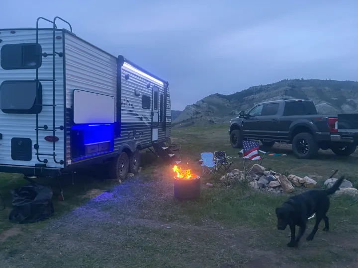 Serene evening at an RV rental campsite. A travel trailer with blue lights, truck, campfire, and black dog under mountain views.. Jayco Jay Flight Baja Edition 2024