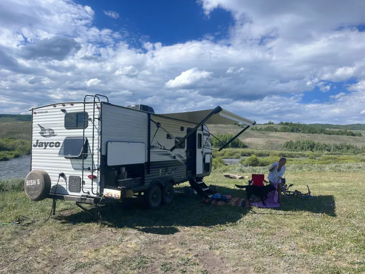 RV rental adventure by a river. A white Jayco trailer with an awning, a person, and a black dog camp in nature under a cloudy sky.. Jayco Jay Flight Baja Edition 2024