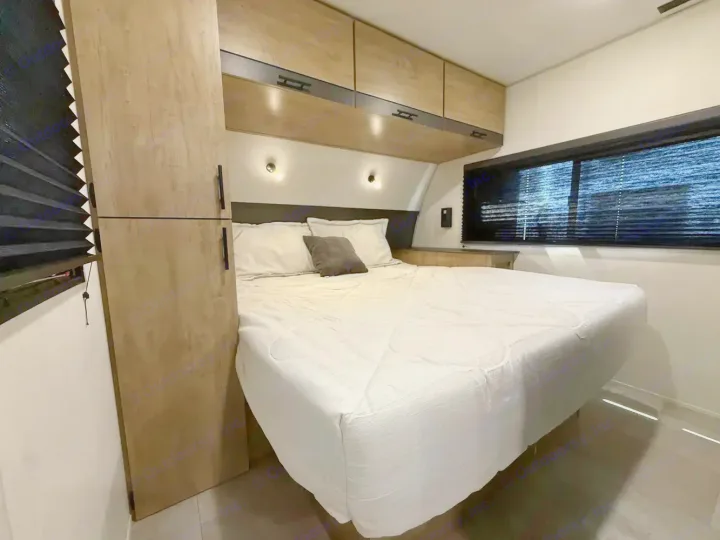 RV rental bedroom: a white-covered bed, wall-mounted lights, overhead storage, and a window with dark blinds.. Forest River Campsite Reserve 2026