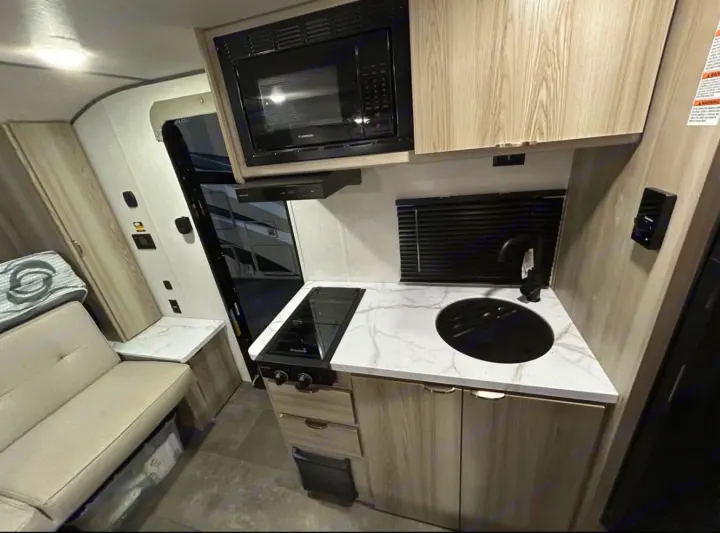 Interior of a modern RV rental, featuring a compact kitchen with a black sink, stovetop, microwave, and light wood cabinets, plus a cozy seating area.. Forest River Salem FSX 2025