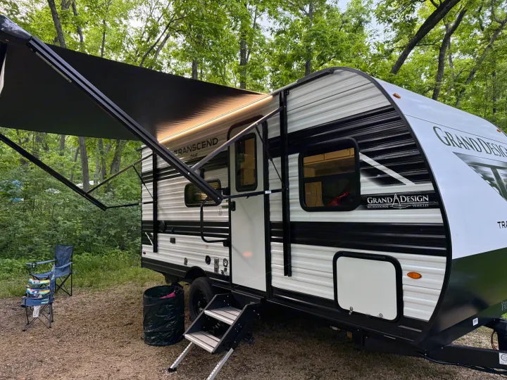 A Grand Design Transcend RV rental is parked at a serene campsite with its awning extended and LED lights glowing.. Transcend One 151BH 2026