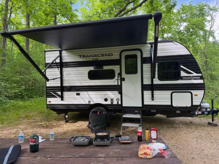 An RV rental parked in a forest campsite with its awning extended. A picnic table in the foreground has outdoor cooking gear.. Transcend One 151BH 2026