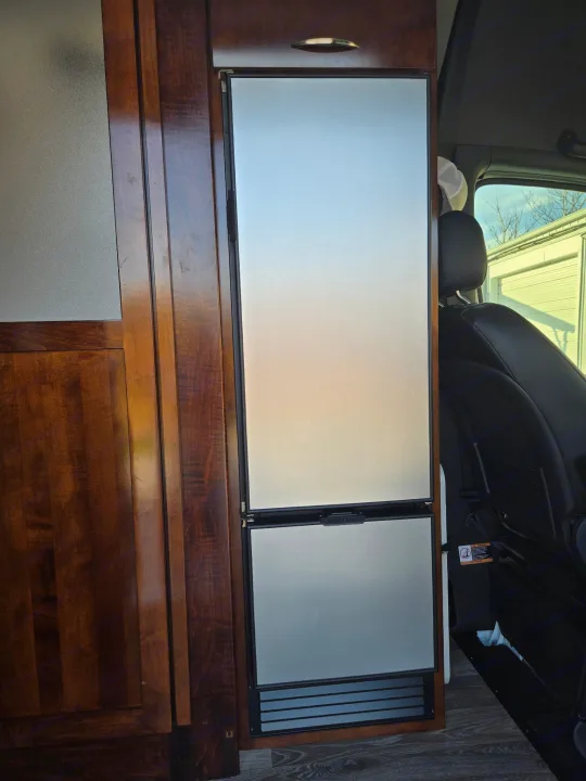 RV rental: sleek, two-door refrigerator/freezer integrated into wooden cabinetry, visible next to a vehicle seat in this camper van.. Coachmen Galleria 2023