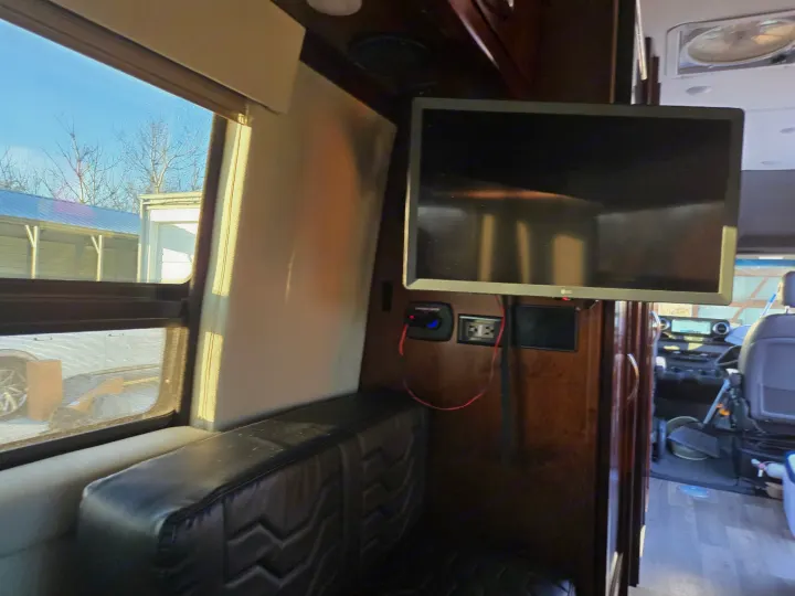 RV rental interior with a mounted TV, black bench seating, and power outlets. A window offers a view of bare trees outside.. Coachmen Galleria 2023