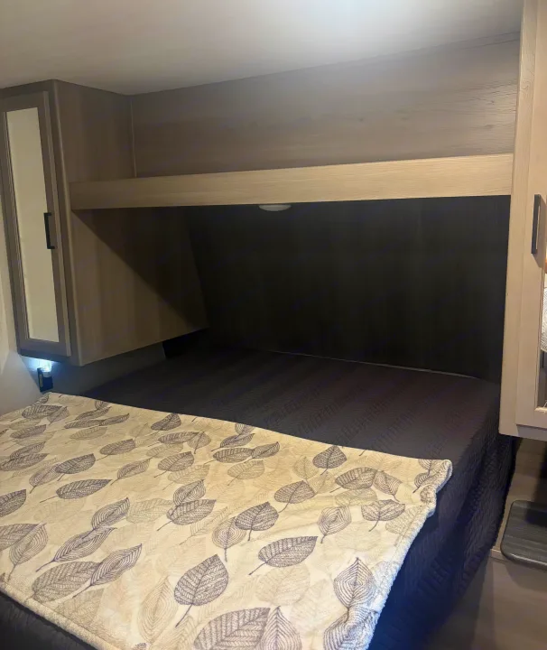 Cozy RV rental bedroom featuring a dark quilted bed, leaf-patterned blanket, and integrated overhead storage with lighting.. Keystone RV Hideout 2024