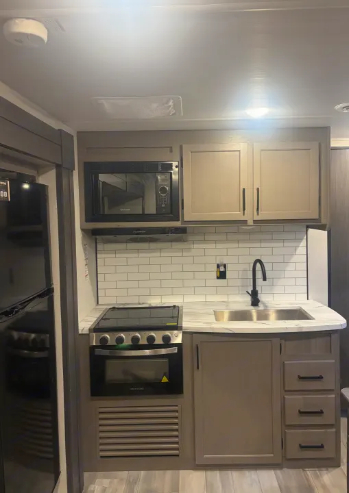 Modern RV rental kitchen featuring microwave, stove, sink, fridge, light wood cabinets, and subway tile backsplash.. Keystone RV Hideout 2024