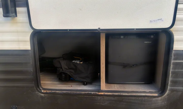 Exterior storage bay of an RV rental, open to reveal a black mini-fridge with a strap and camping gear in bags.. Keystone RV Hideout 2024
