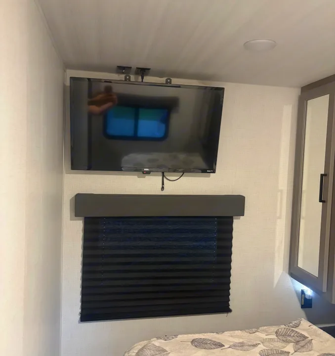 Inside an RV rental, a compact bedroom shows a wall-mounted TV above a window with a dark pleated blind and soft bedding.. Keystone RV Hideout 2024