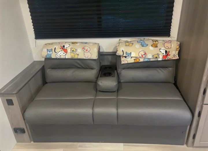 RV rental interior featuring a grey sofa with cupholders, topped with dog-patterned blankets, under a black blind.. Keystone RV Hideout 2024