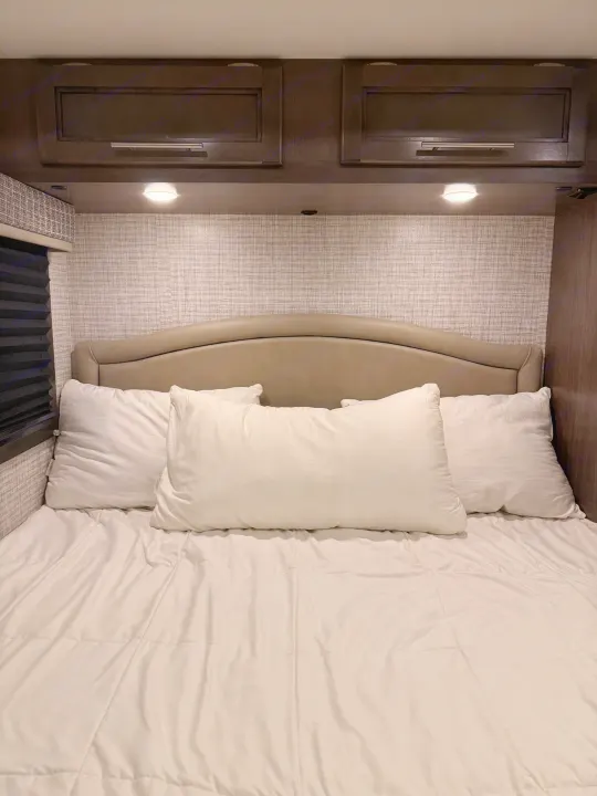 Cozy RV rental bedroom featuring a bed with white pillows and comforter, dark wood cabinets, and task lighting.. Jayco Melbourne 2024