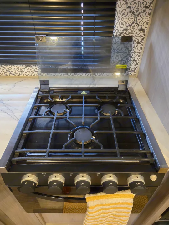 RV rental kitchen cooktop: Four-burner gas stove with black control knobs and a clear glass cover, shown against blinds.. Dutchmen Coleman Lantern 2022