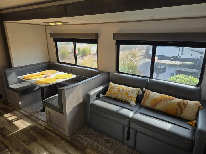 Inside an RV rental: a cozy dinette with a table and a comfortable sofa with decorative pillows, surrounded by large windows.. Dutchmen Coleman Lantern 2022