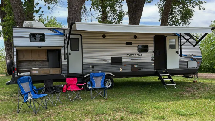 This rv rental, a travel trailer with an open outdoor kitchen, extended awning, and camping chairs, sits in a grassy area.. Coachmen Catalina Legacy 2022