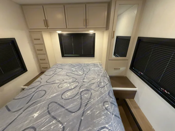 RV rental bedroom featuring a mattress in plastic, overhead cabinets, side windows with blinds, mirror, & drawers.. 2027
