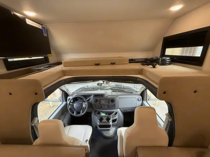 RV rental interior: Cab-over bunk, TV, and cockpit dashboard from the living area. Shows front seats and steering wheel.. 2027