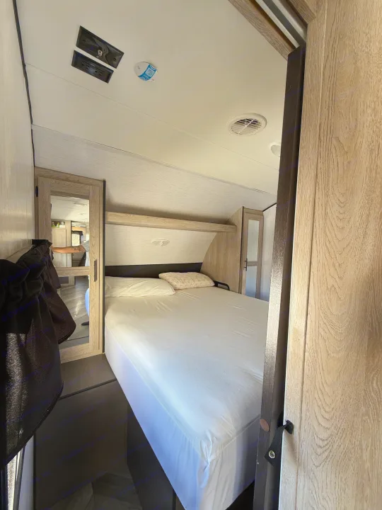 Inside an RV rental: a bright and cozy bedroom featuring a white bed, wood-paneled walls, and compact storage.. Heartland RVs Prowler 2022