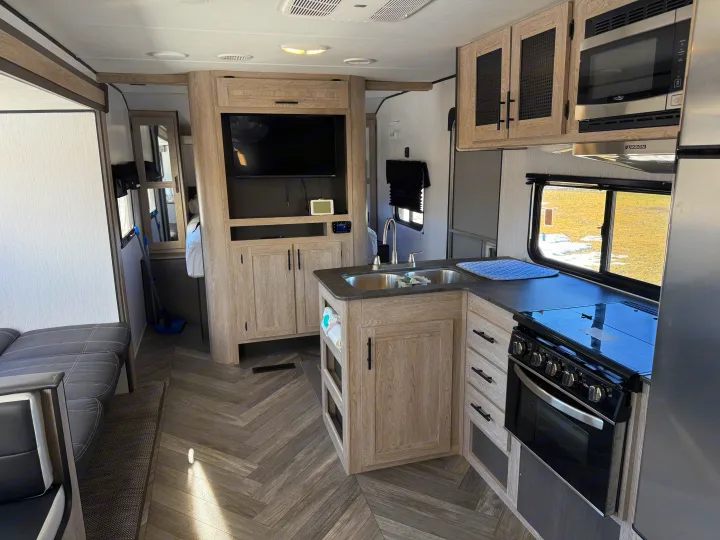 This modern RV rental interior features a kitchen with stove, microwave, and sink, a comfy living area, and stylish herringbone floors.. Heartland RVs Prowler 2022